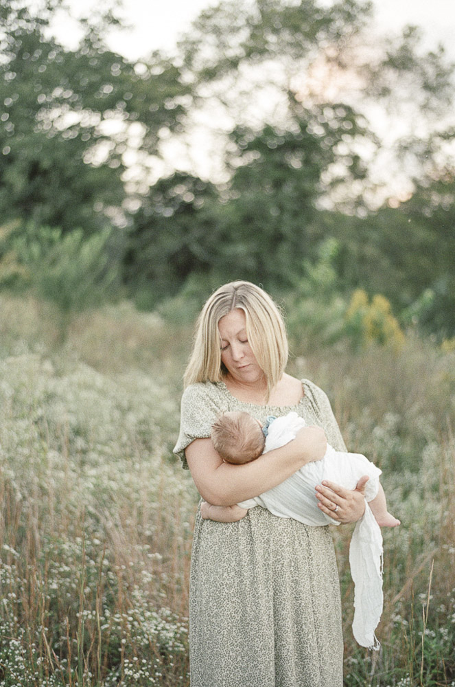 A young mother holds her infant in white swaddling cloth in a field at sunset