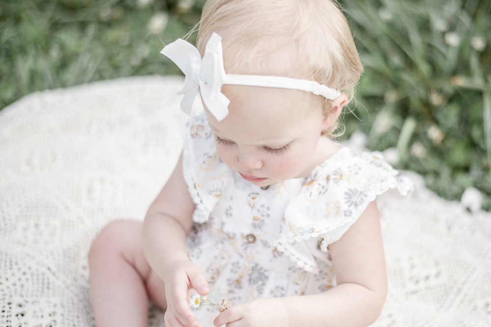 A baby with long eyelashes looks a flower she holds in her hand.