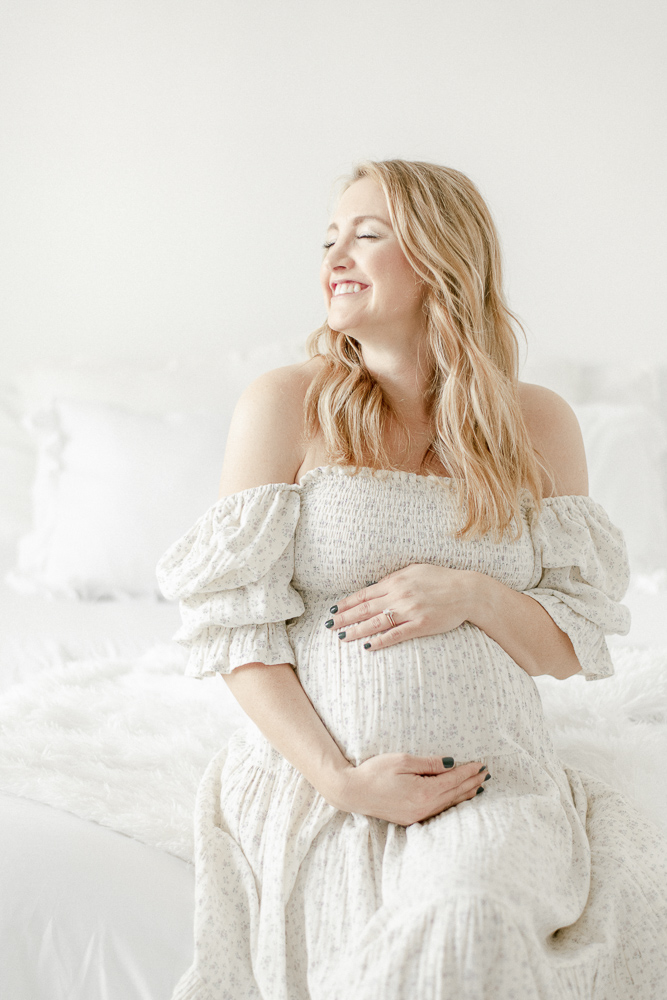 A pregnant mother in a white dress smiles with her eye closed as she hugs her belly