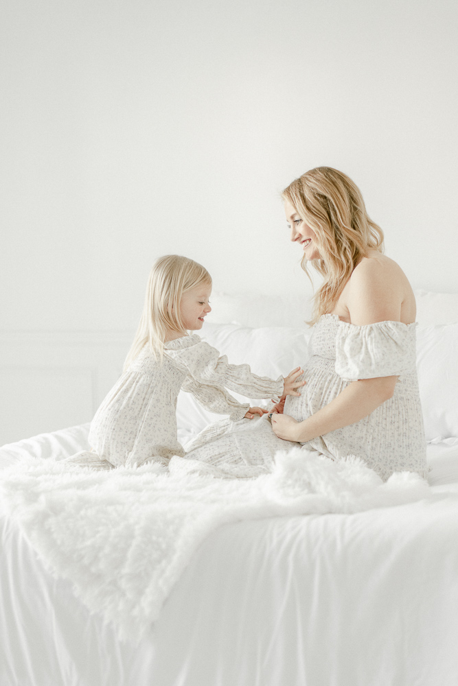 A blonde toddler touches her mother's pregnant stomach as they sit on a bed