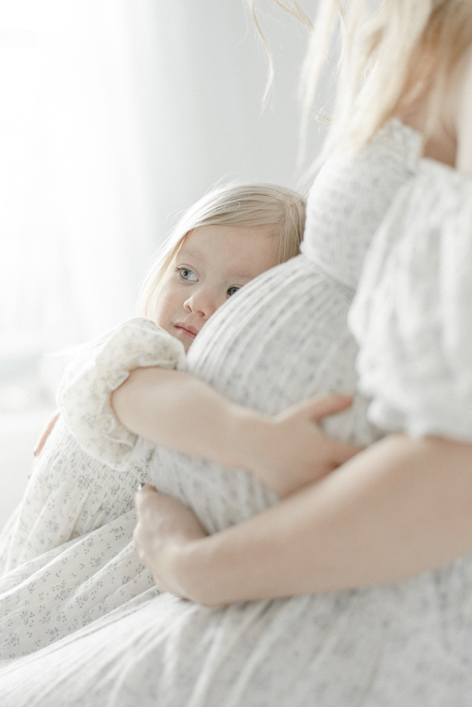 A blonde toddler girl hugs her mother's pregnant stomach.