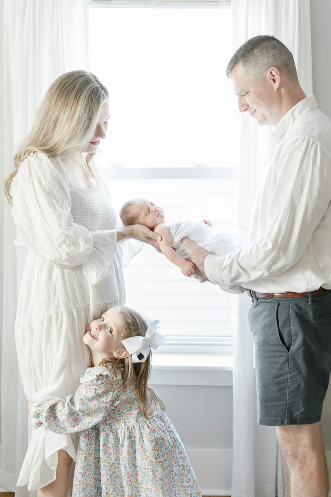 A mother and father hold a newborn baby in front of a window