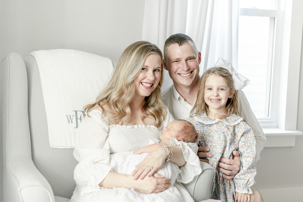 A young family of four smiles with a newborn and young toddler girl during in-home newborn photos