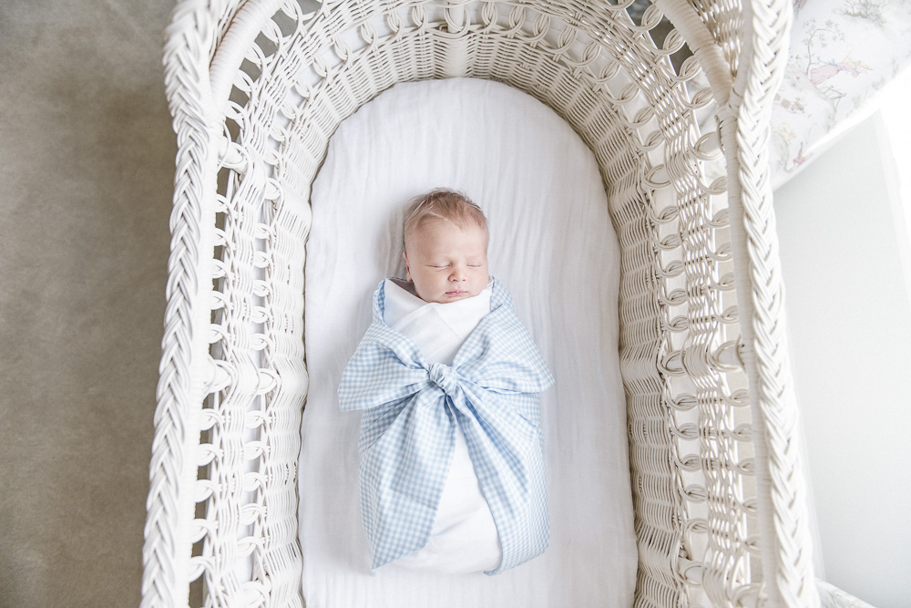 Newborn baby boy in a Beaufort Bonnet swaddles sleeping in a white bassinet .
