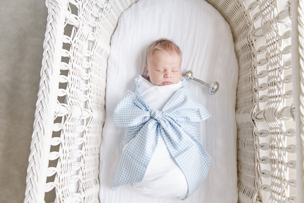 Newborn baby boy in a Beaufort Bonnet swaddles sleeping in a white bassinet with a silver rattle.