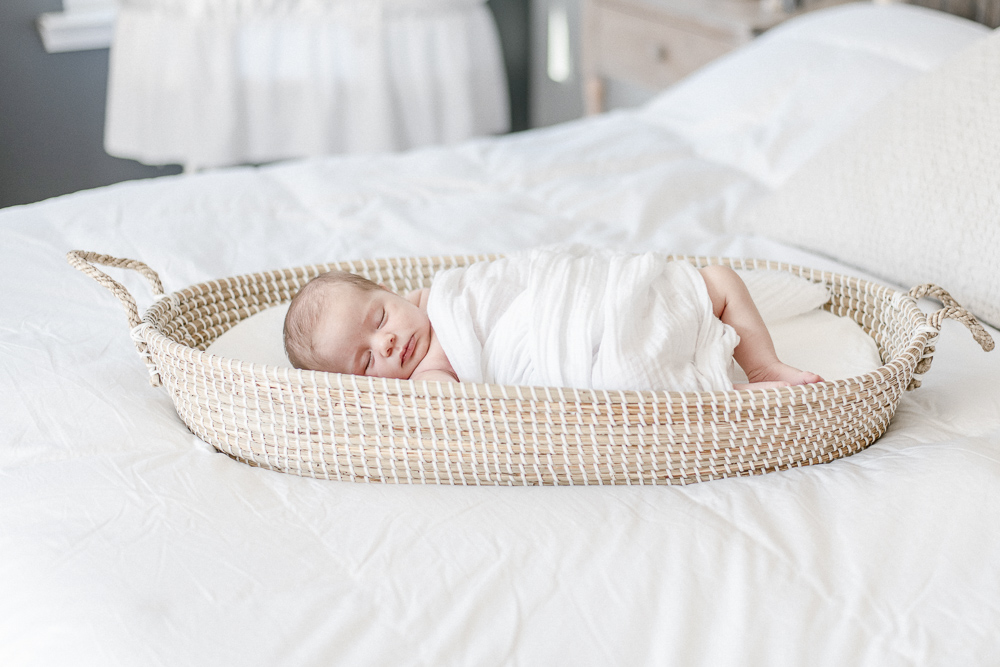 A newborn swaddled in white cloth sleeps in a brown wicker basket