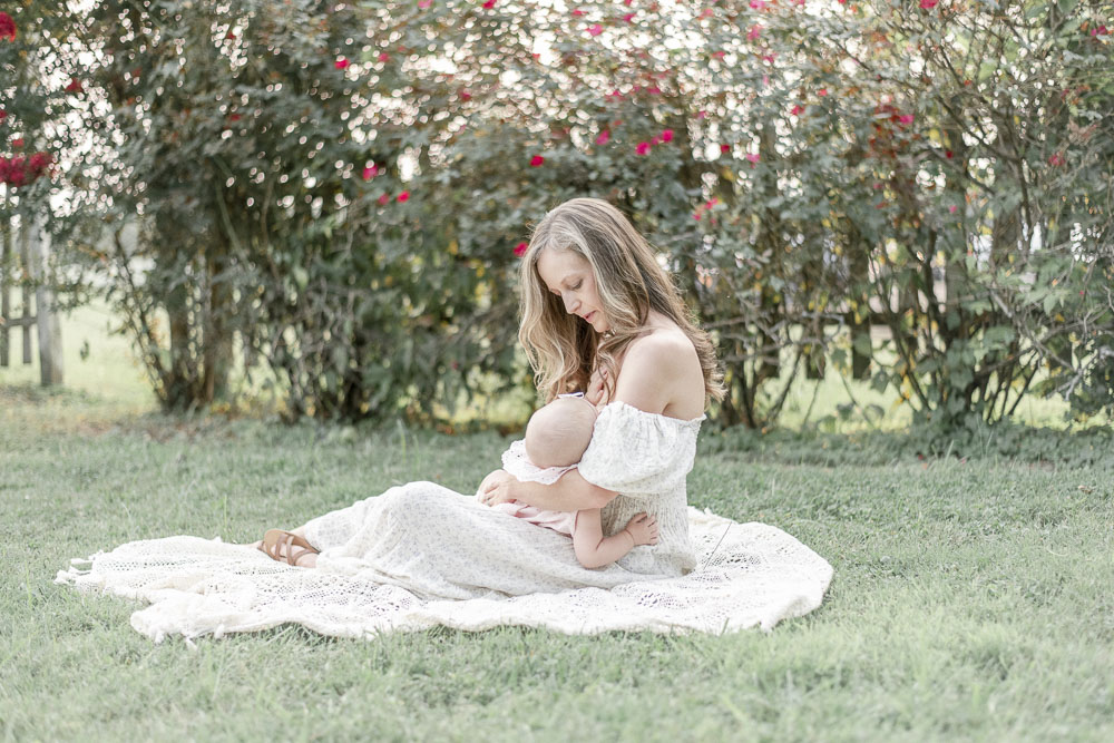 A mother sits on a blanket in the grass and breastfeeds her baby girl.