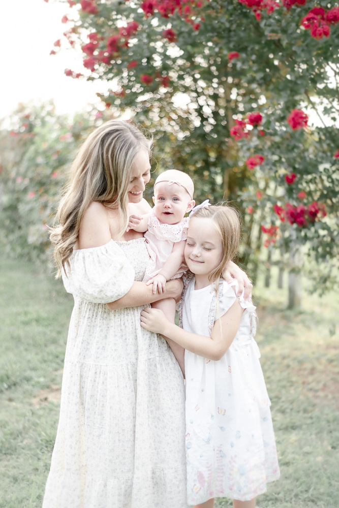 A mother hugs her two daughters in front of pink flowering trees.
