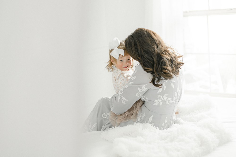 A mother nuzzles her toddler daughter as this sit together on a white bed