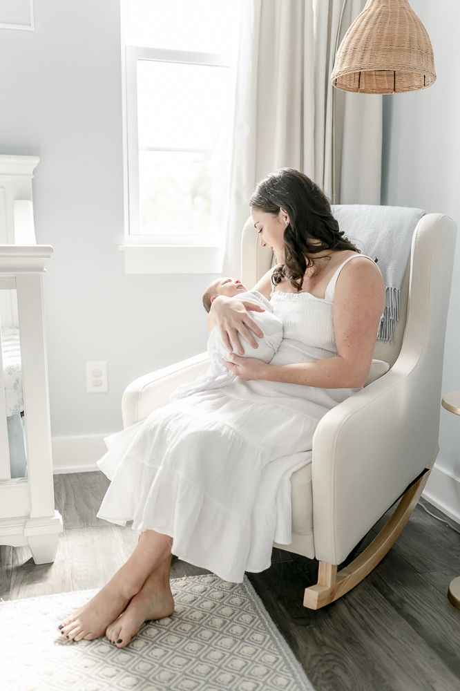 A brunette mother holds her baby in a white rocking chair near a window in a blue nursery by the best nashville newborn photographer Kristie Lloyd