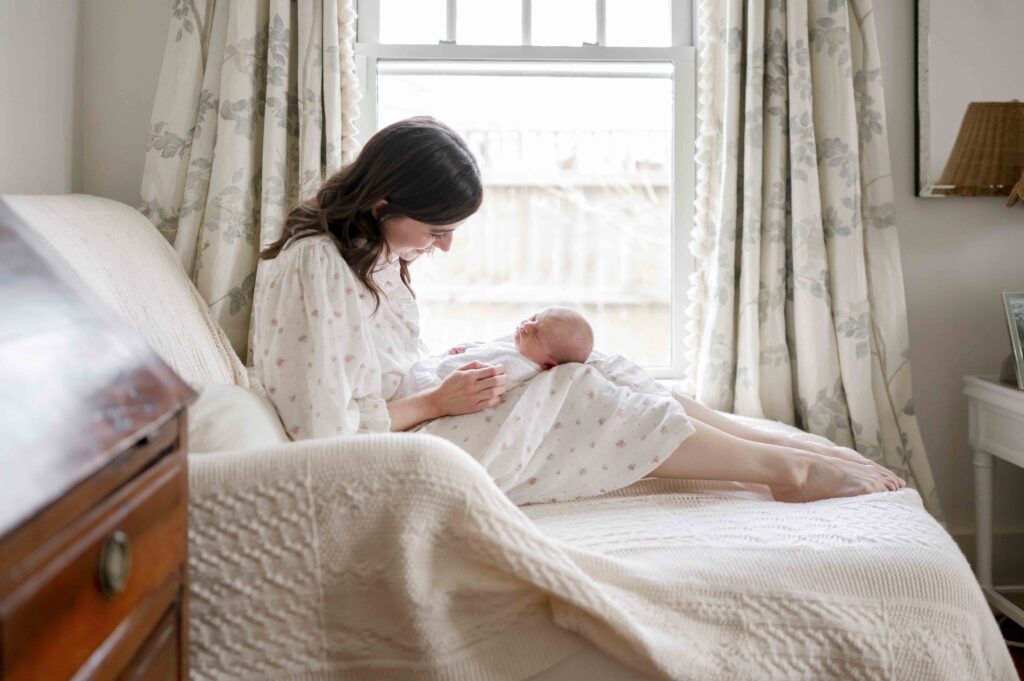 A mother sits in a chair by a window and smiles at her newborn baby