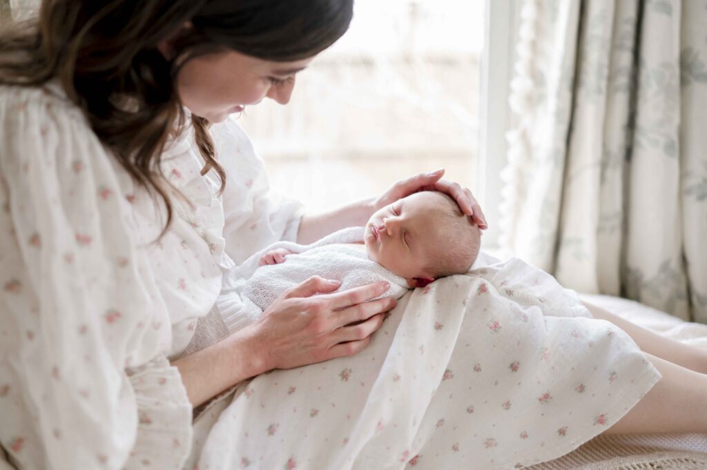 A mother sits in a chair by a window and smiles at her newborn baby