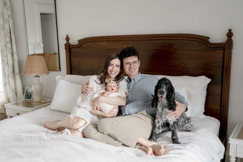a mother and father hold their newborn baby on the master bed while their dog sits beside them