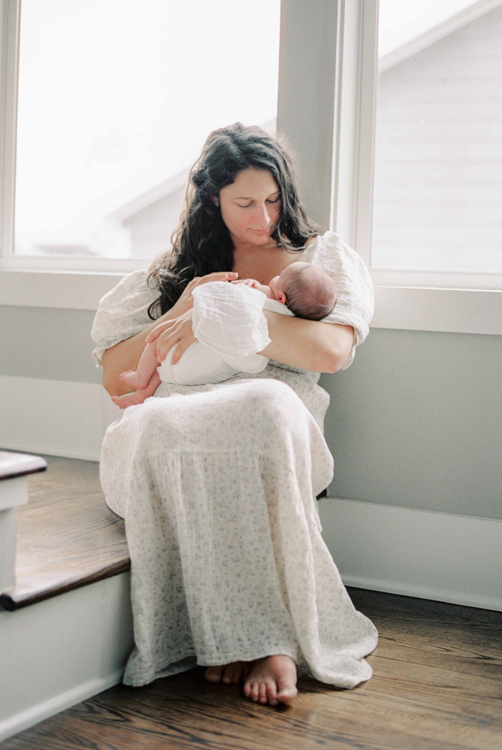 A mother cradles her baby sitting in front of a window