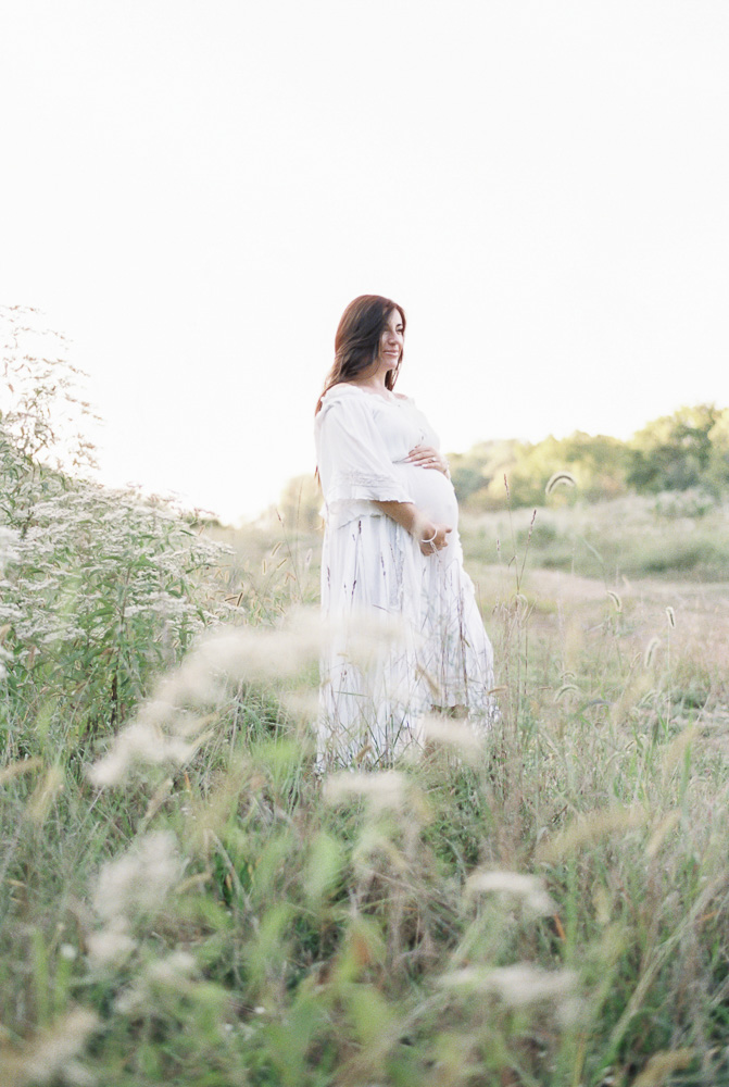 A pregnant woman in a white dress poses in a field of wildflowers