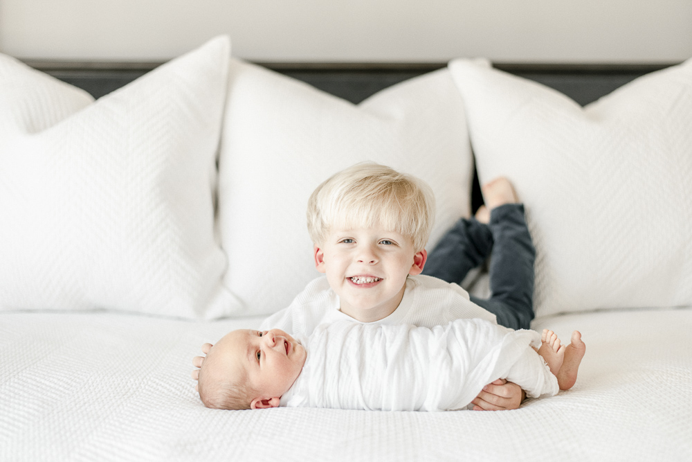 A toddler and newborn lay together smiling on a bed with white pillows