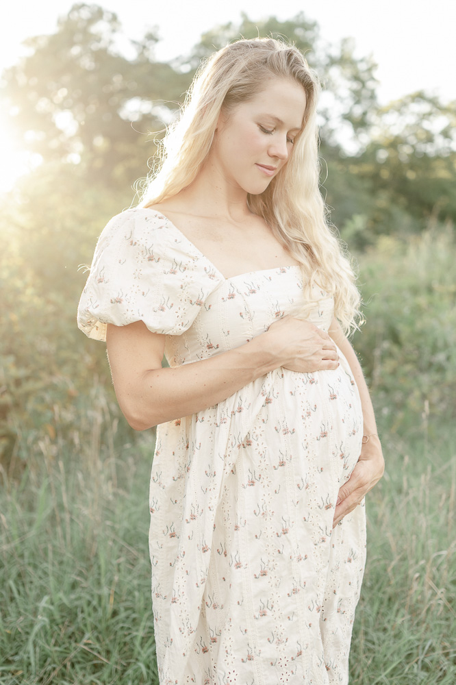 A pregnant woman smiles down at her stomach with a golden sun flare shining over her shoulder