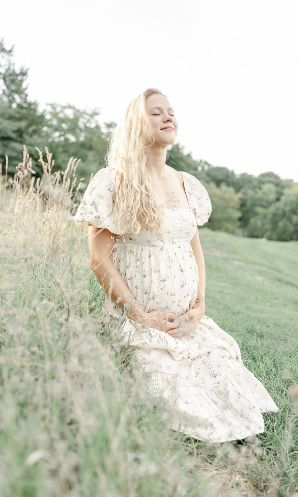 A pregnant woman smiles joyfully holding her stomach as she kneels on a middle tennessee hillside 