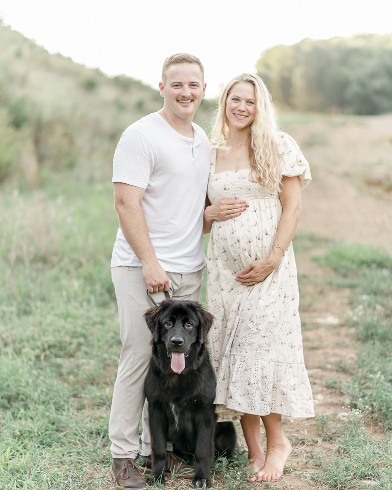 Pregnant couple and their black Labrador stand on a hill at sunset
