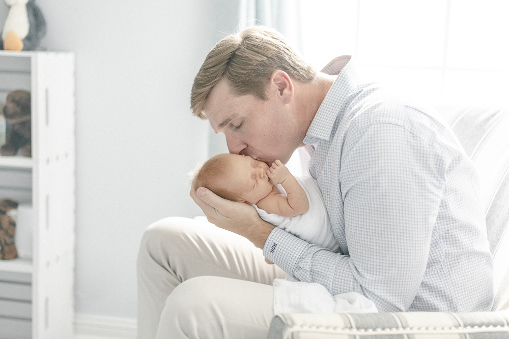 A father sits in a chair in his Nashville home kissing his newborn baby on the cheek
