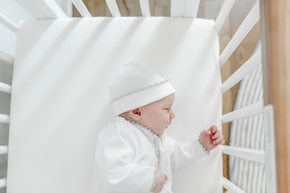A newborn baby boy sleeps in his crib wearing a feltman brothers gown and hat in his Nashville home's nursery.