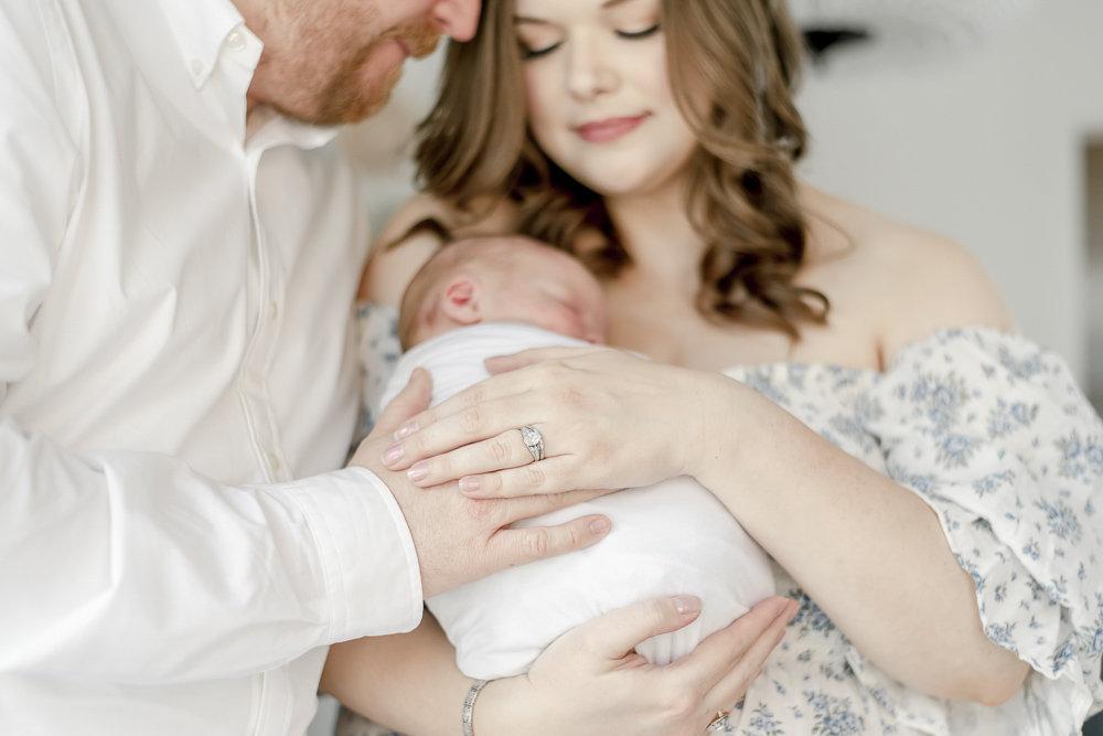 New parents hold their newborn baby, an emphasis on their hands overlapping and the mother's diamond wedding ring