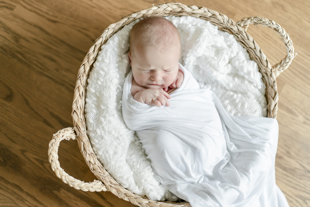 A newborn wrapped in white sleeps in a basket on a white blanket