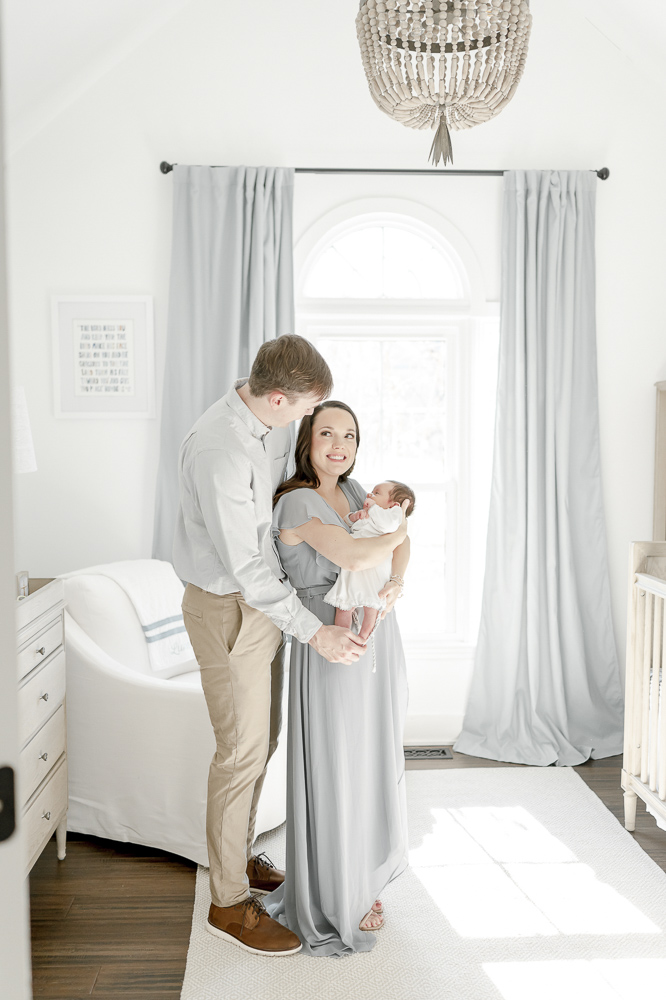 A married couple holds their baby boy standing in front of a large nursery window with blue curtains