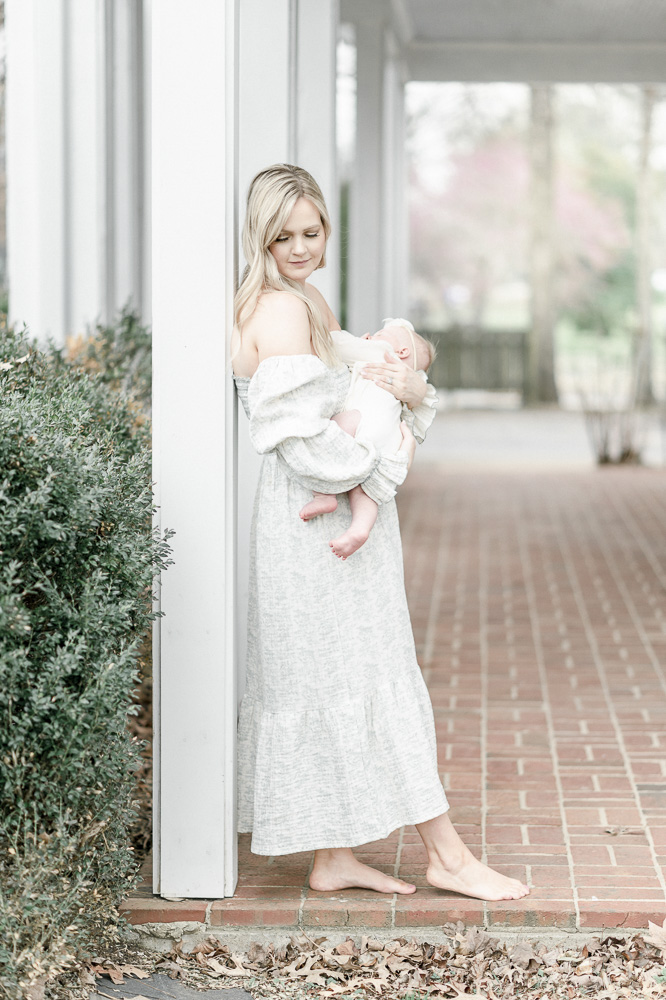 A mother in a white dress stands on a white porch in downtown Franklin TN holding her baby