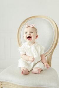 6 month photos of baby girl in a pink smocked dress sits in a chair smiling in Nashville baby photographer Kristie Lloyd's studio