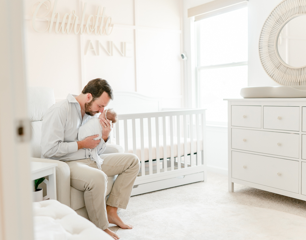 A father sits in the nursery rocking chair and kisses his newborn  by Brentwood newborn photographer Kristie Lloyd.