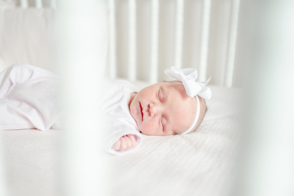 Newborn baby girl sleeps in her crib on pink gingham sheets wearing a white bow for her in-home session with Kristie Lloyd