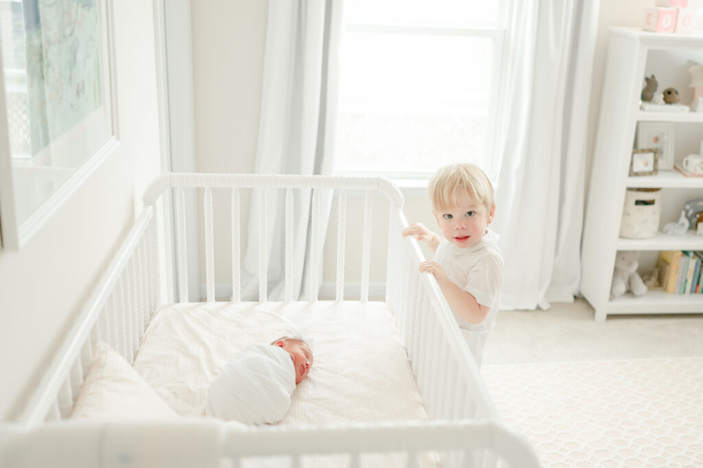 A toddler boy stands on his newborn baby sister's crib 