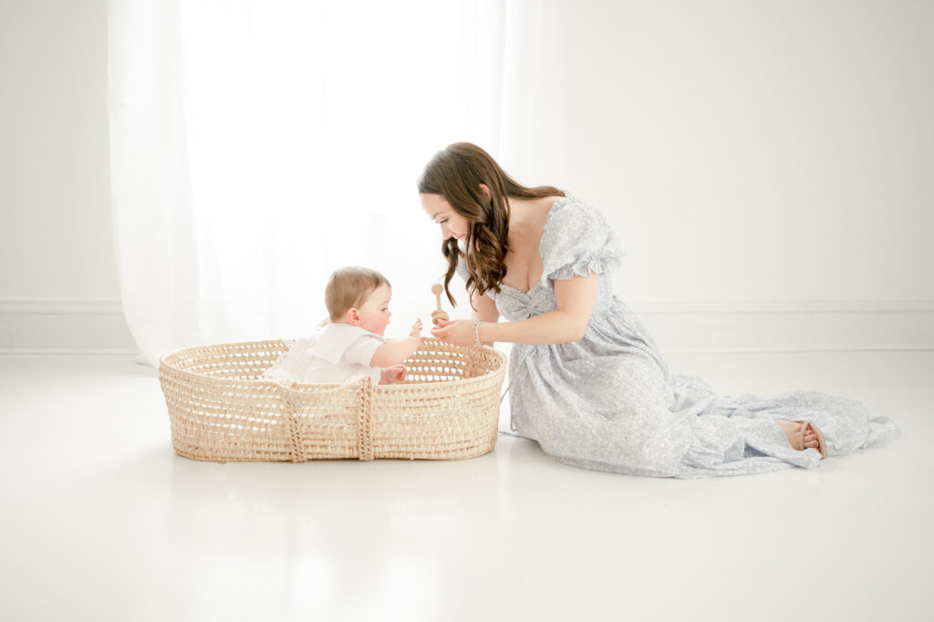 A mom holds a rattle to show her baby sitting in a basket at Kristie Lloyd's studio for his 6 month photoshoot 