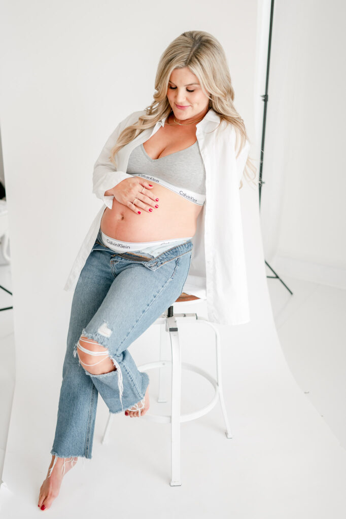 Pregnant woman in jeans and calvin klein underwear sits on a stool during her studio maternity photos with Kristie Lloyd. 