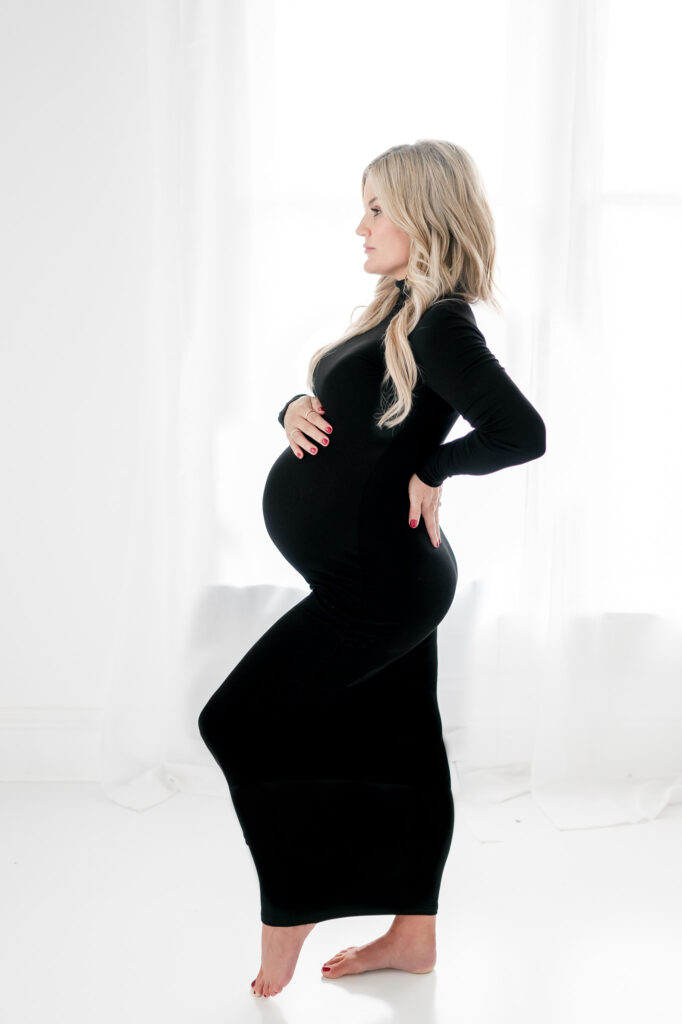 Pregnant woman in black fitted dress stands during her studio maternity photos with Kristie Lloyd. 