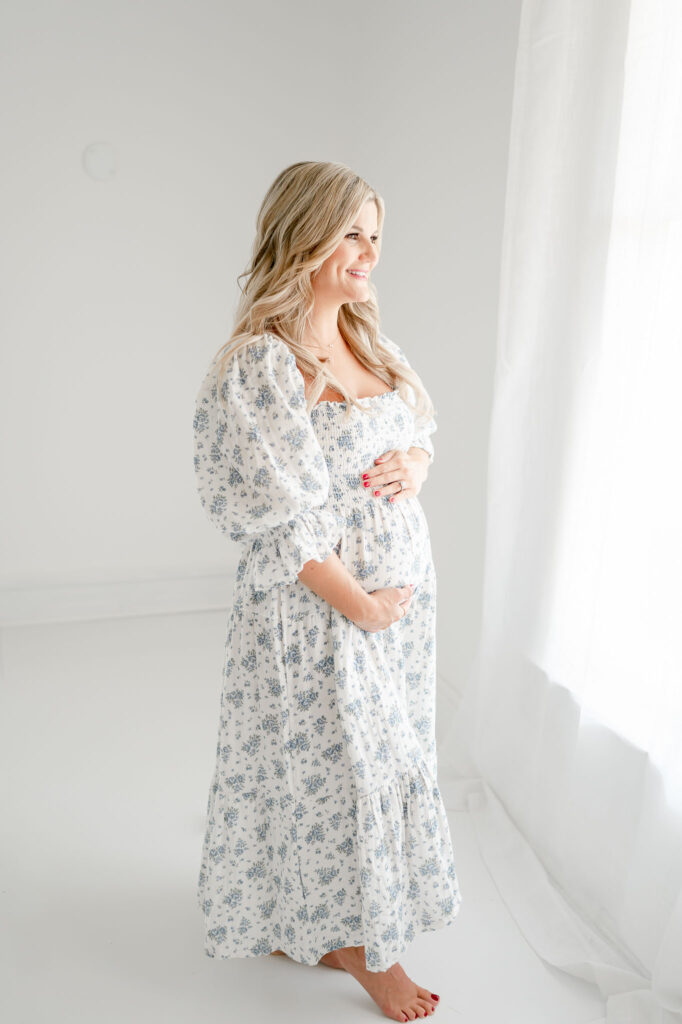 Pregnant mother in blue floral dress smiles out a window during her studio maternity photos.