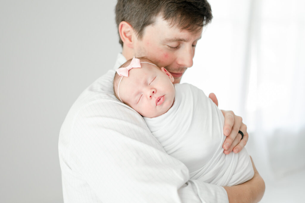 Father holds his newborn baby wearing a white swaddle and pink bow