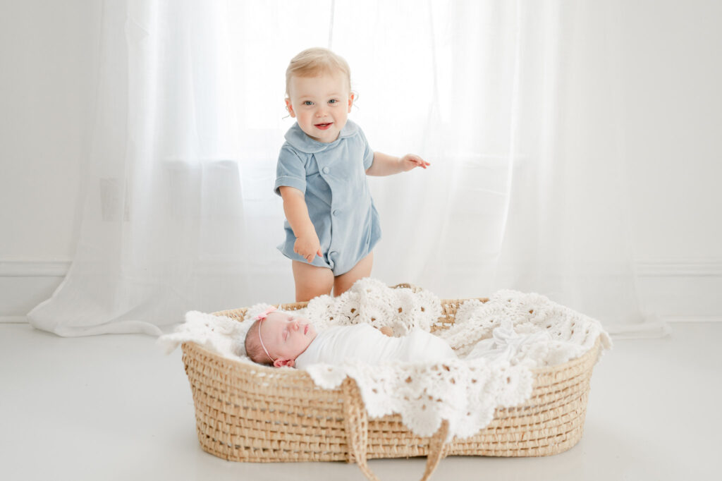 Toddle sibling stands over his newborn sister during studio newborn session