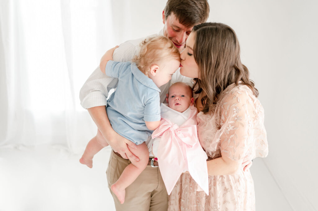 Mother kisses her toddler son while holding her newborn baby girl during their studio newborn session