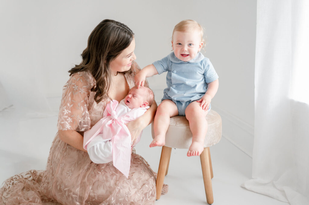 Toddler boy points at his baby sister that is held by mom