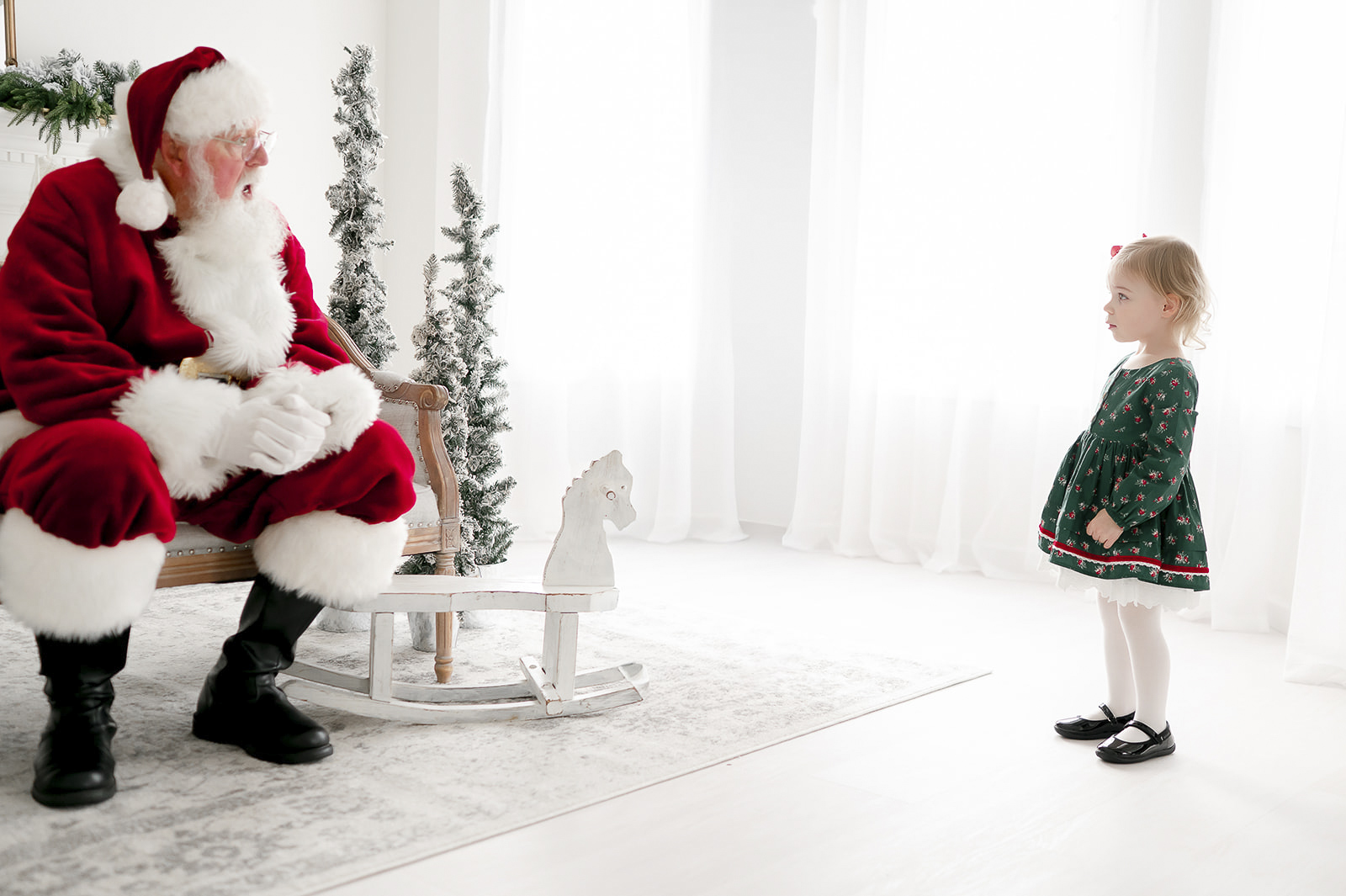 A toddler girl stands far away as she looks at Santa sitting on a bench in Kristie Lloyd's studio