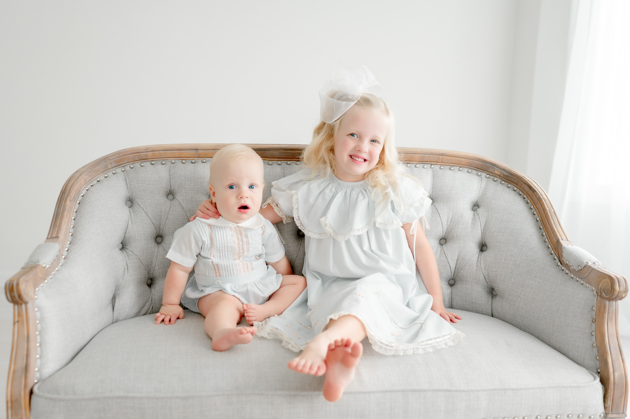 A baby boy and his toddler sister sit on a gray tufted bench in blue heirloom outfits