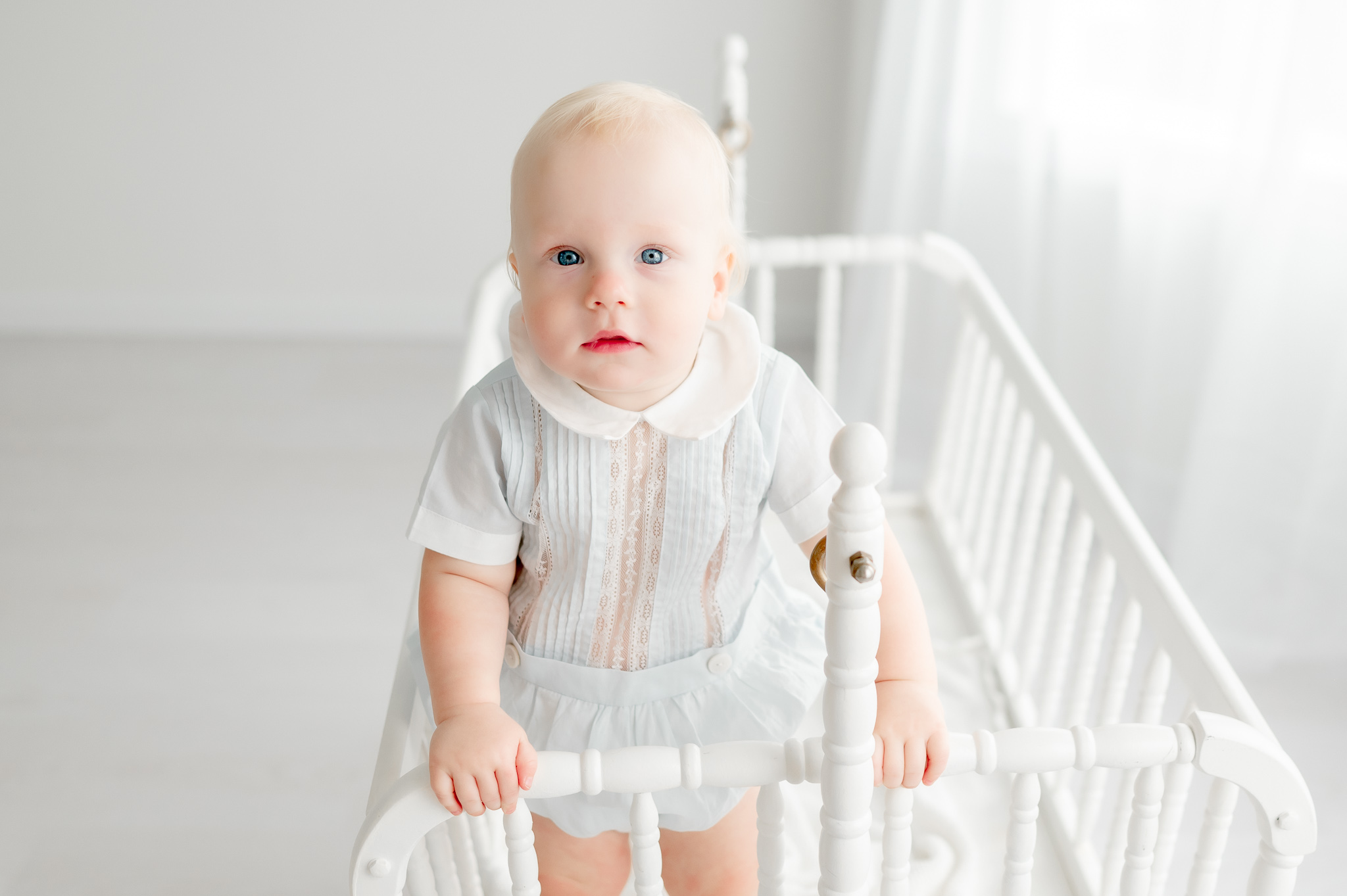 Baby boy stands in a white jenny lind crib in Kristie Lloyd's photography studio wearing an heirloom outfit