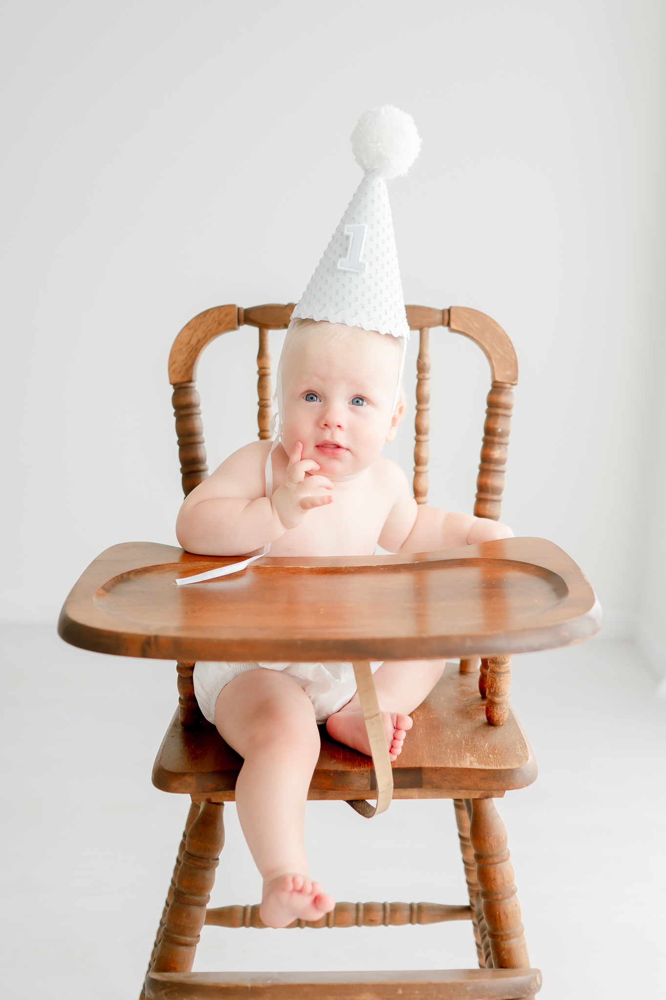 Baby boy sits in brown high chair with a party hat