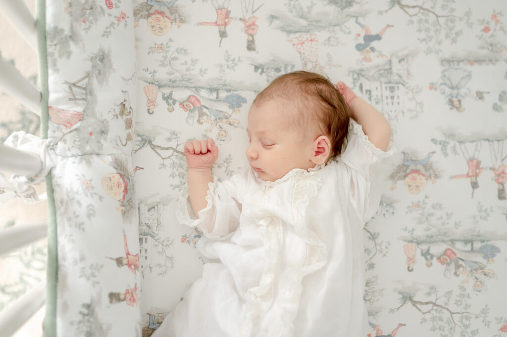 A sleeping newborn lays in a vintage Feltman brothers gown with a toile pattern on his crib sheet