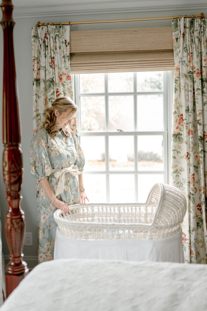 A mother stands next to a window smiling down into a white wicker bassinet 