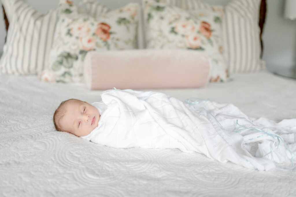 A baby boy lays swaddled on a bed with floral pillows behind him