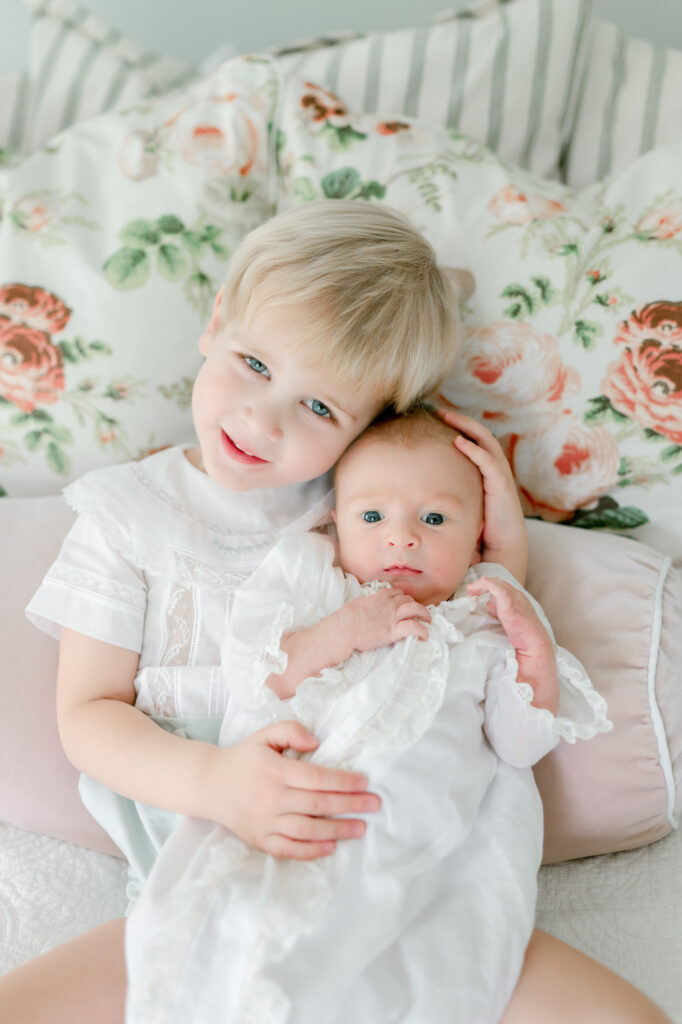 A blonde toddler boy holds his newborn brother sitting against floral pillows