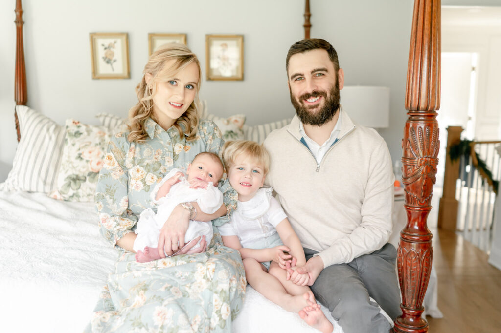 A family of four sits on a 4 post bed smiling as they hold a newborn